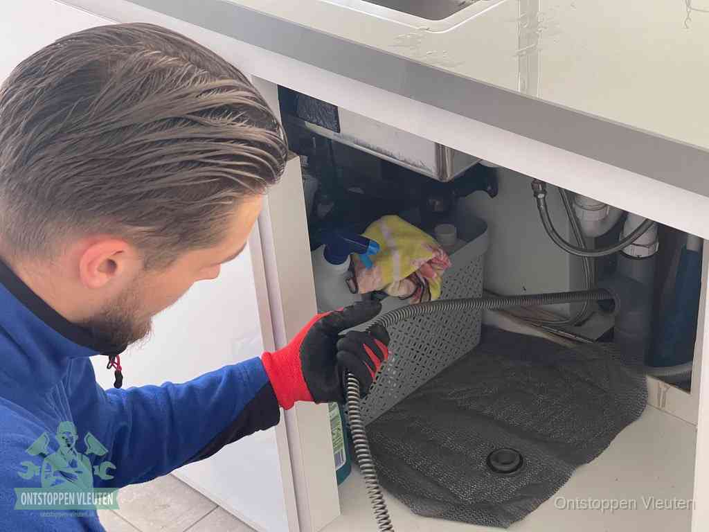 Een monteur voert een flexibele ontstoppingsveer in onder het keukenkastje, langs de sifon, om de gootsteen te ontstoppen in Vleuten