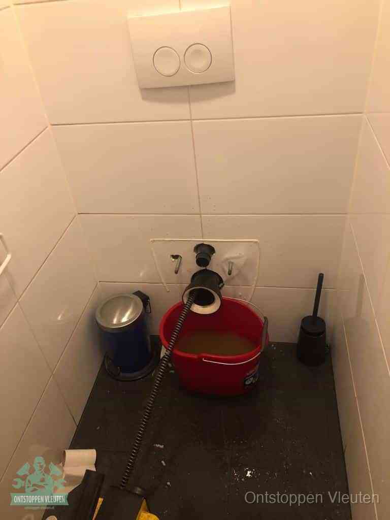 Een ontstopte wand-wc met verwijderde toiletpot waar een ontstoppingsveer in de wanduitgang verdwijnt, het achterhalen waar de verstopping zit in Vleuten