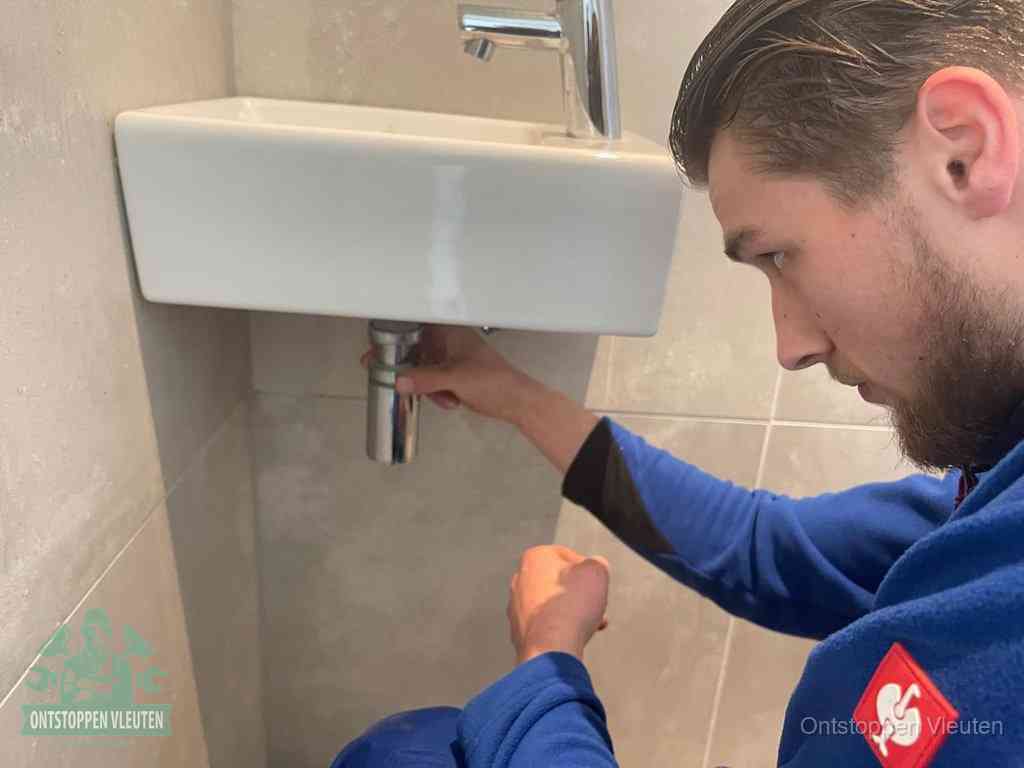 Een monteur draait met de hand de zwanenhalssifon onder een fonteinwasbak in de toiletruimte los om de bron van rioollucht in Vleuten te vinden